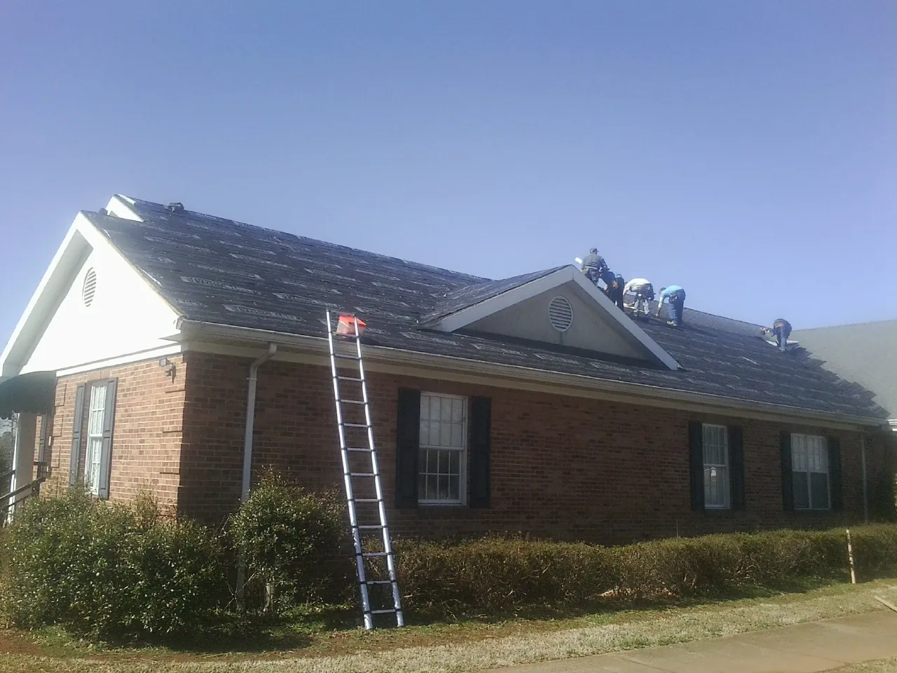 Crew tearing shingles off a brick ranch roof with a ladder set — Emergency Roof Repair work by our crew in Garrattsville