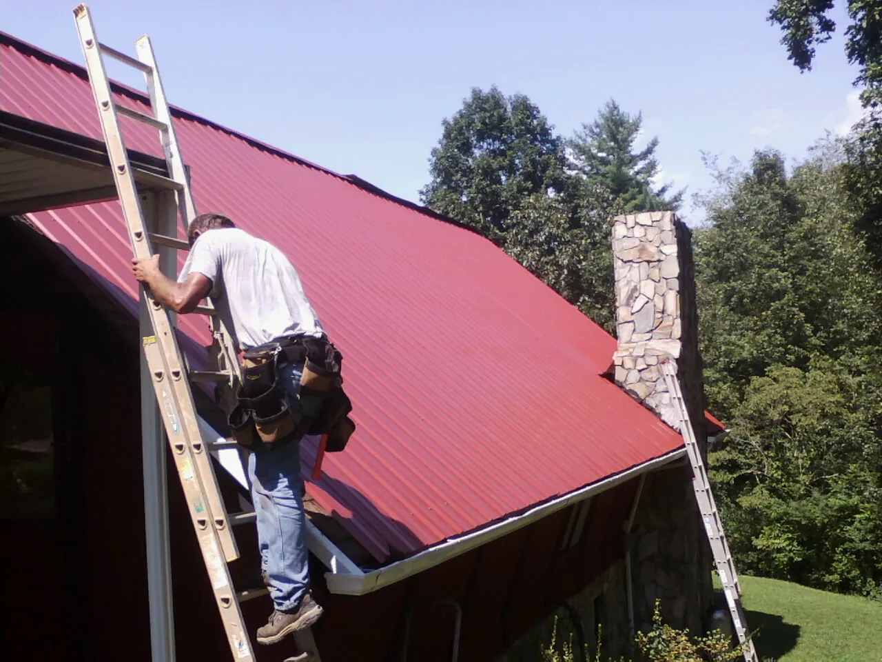 Roofer on a ladder beside a red metal roof and stone chimney — Soffit & Fascia Repair by Roofing Brothers in Garrattsville